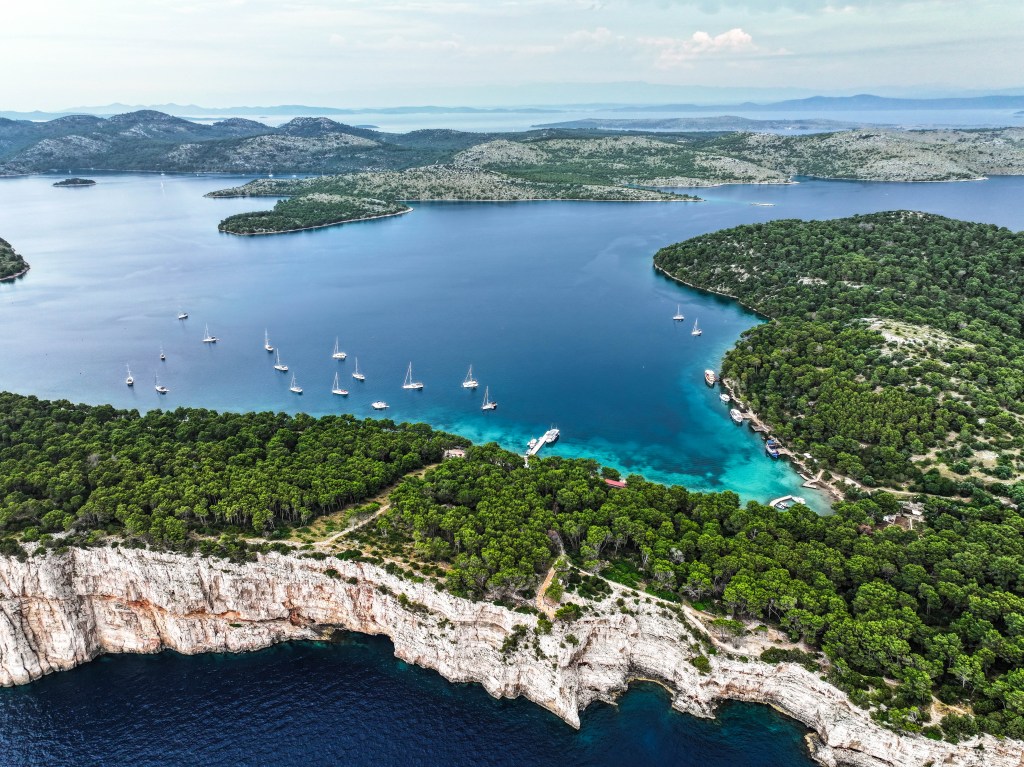 Scenic aerial view of Telašćica Nature Park with dramatic cliffs, saltwater lake, and anchored sailboats surrounded by lush forest and Adriatic Sea.