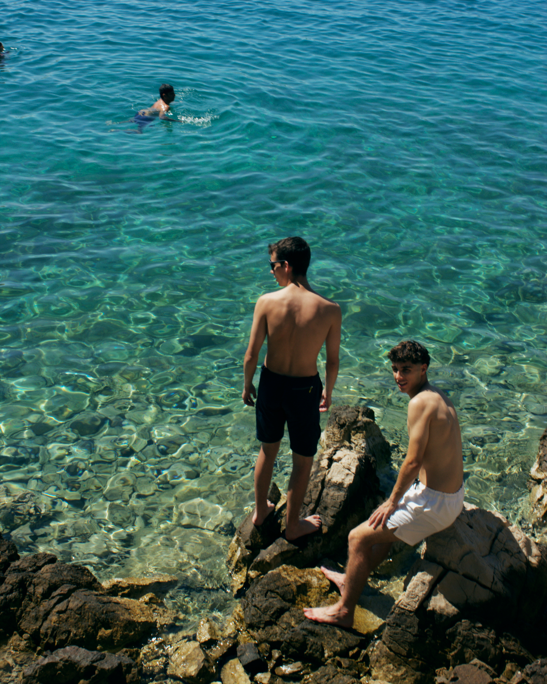 Two men sitting on rocks above crystal-clear turquoise water near Kornati islands during a swimming stop.